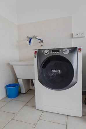 Eine Waschmaschine mit Frontlader, ein blaues Eimer und ein weißes Waschbecken in einem Badezimmer.