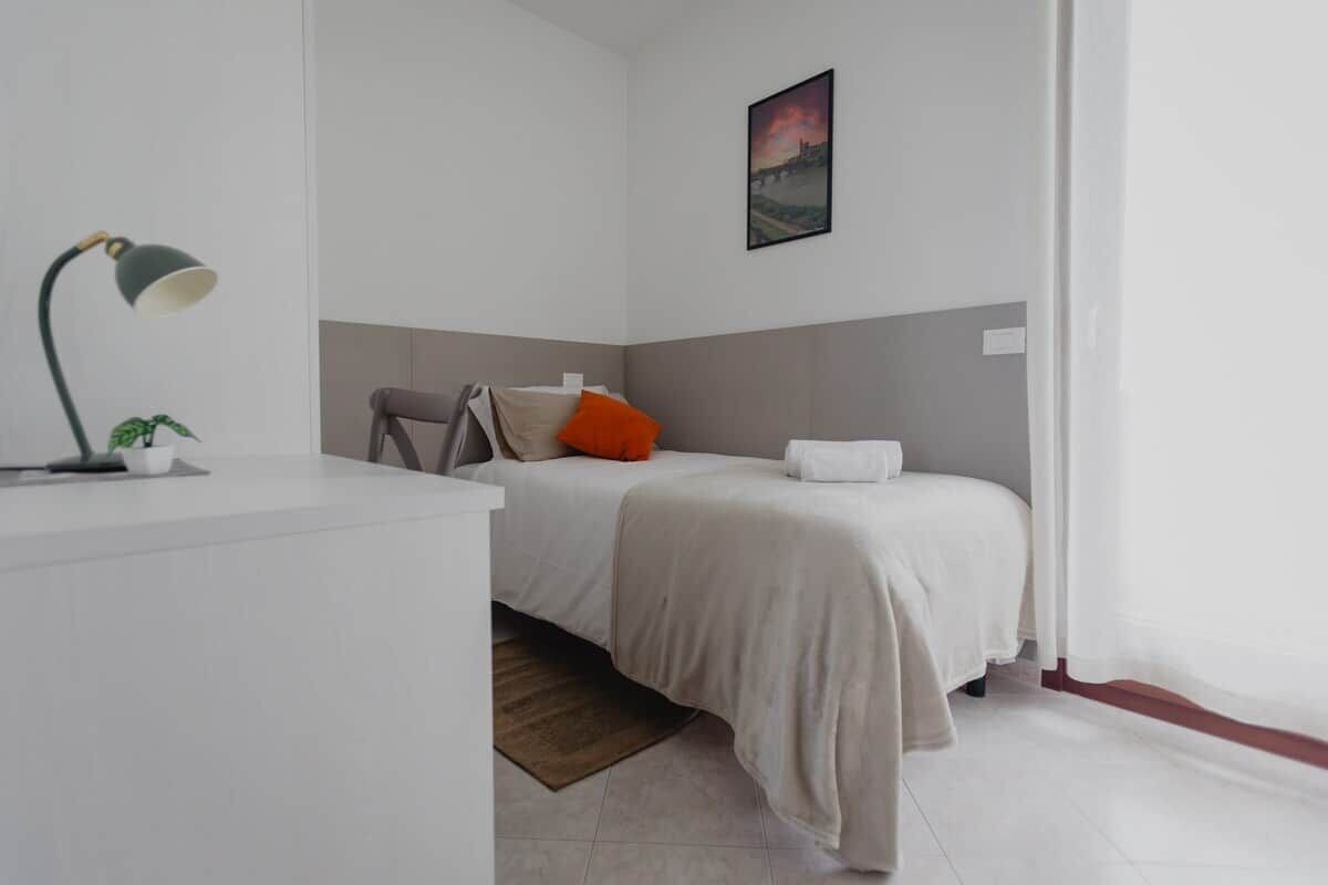 A minimalist bedroom with a bed, a desk, a lamp, and a single potted plant.