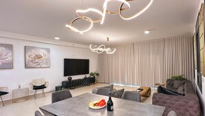 A modern living room with a dining area, featuring a sofa, a TV, and a table with a bottle and fruit. - Sky26 Mini penthouse with 6-Rooms Sew View Bat Yam (Bat Yam)