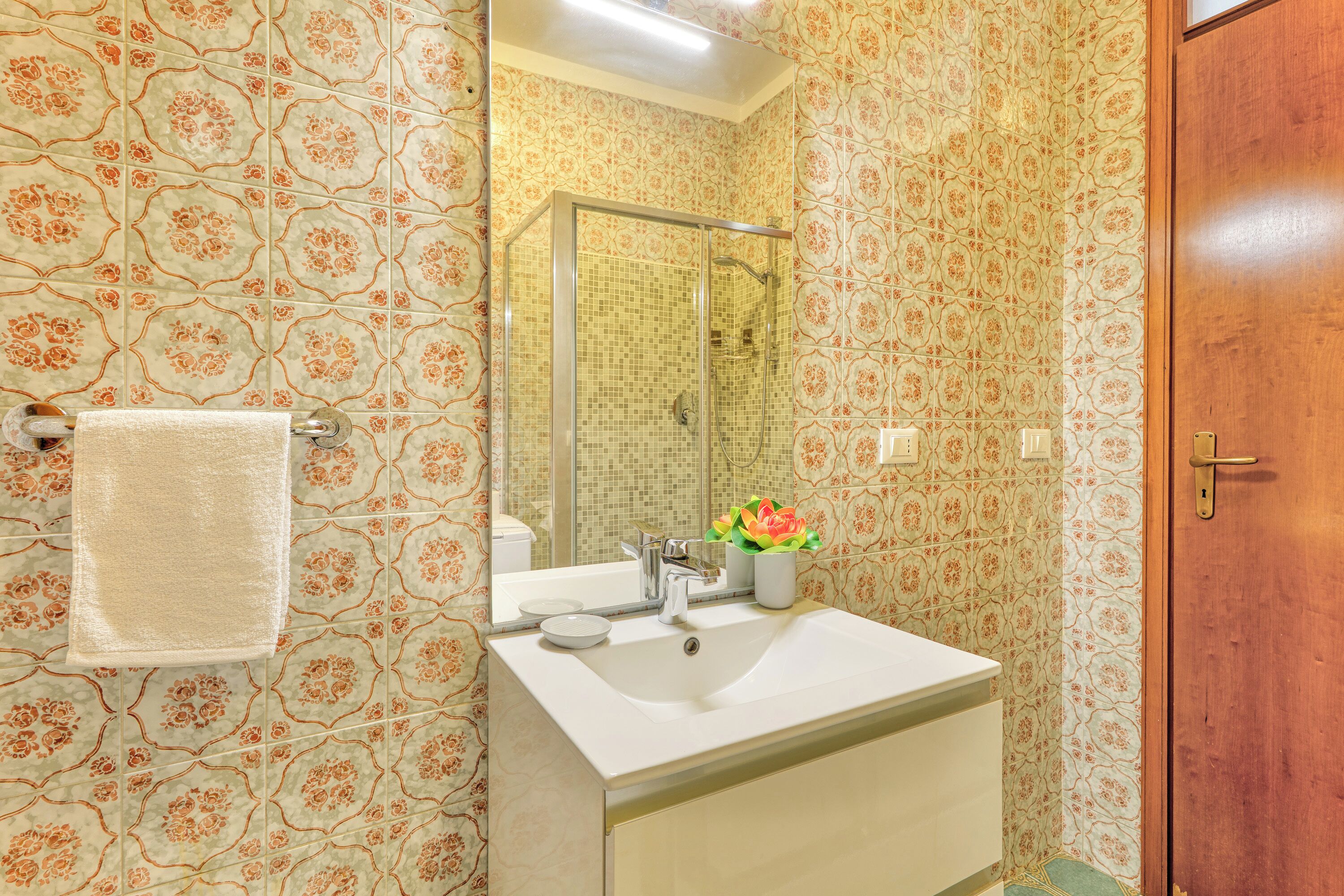 Ein Badezimmer mit blumigem Wandpapier, einem weißen Waschbecken, einer Duschkabine aus Glas und einer Holztür.