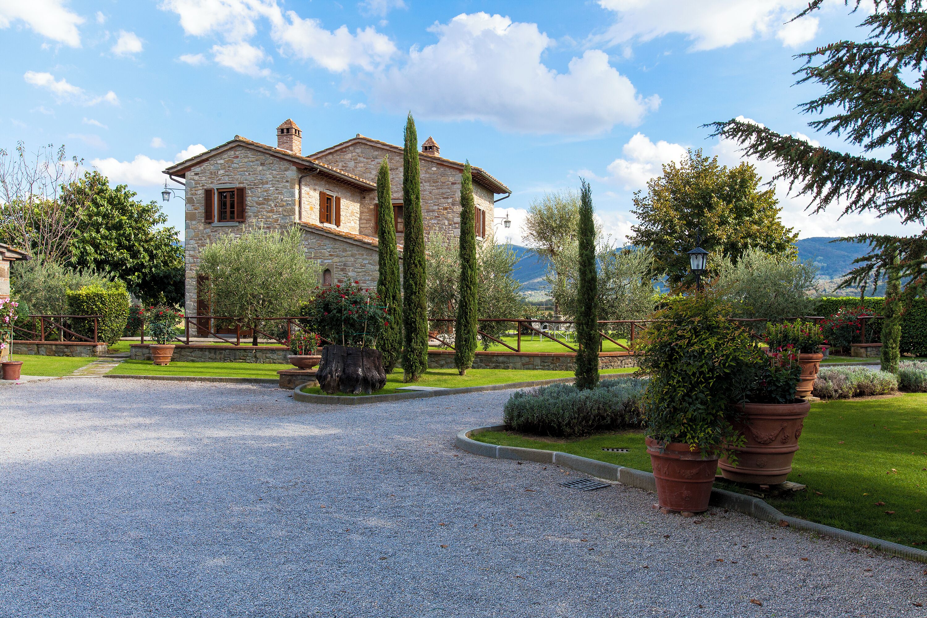Une maison en pierre avec une allée gravillonnée, entourée de jardins bien entretenus et de grands arbres.