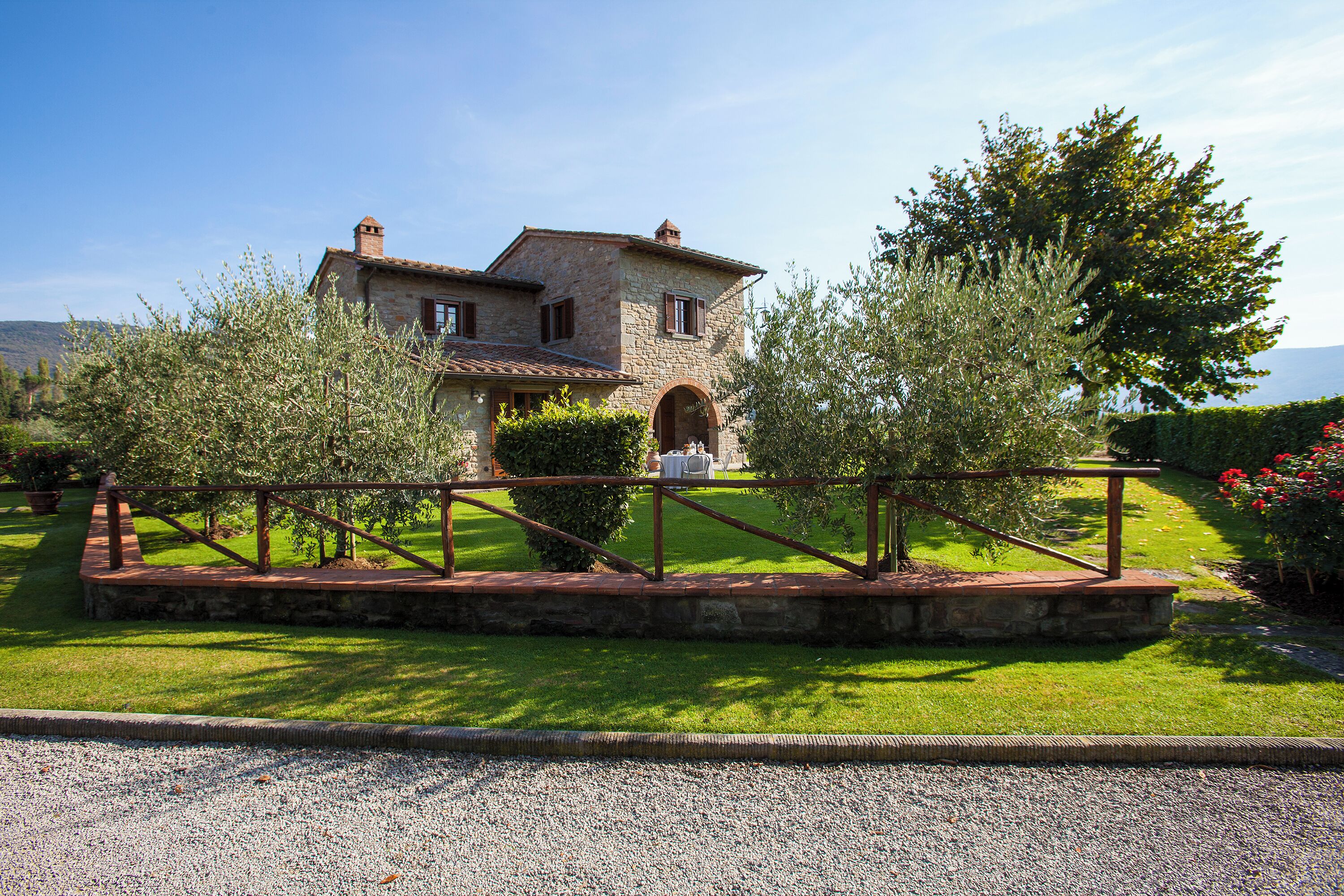 A stone house with a tiled roof, surrounded by a gravel driveway and a metal fence.