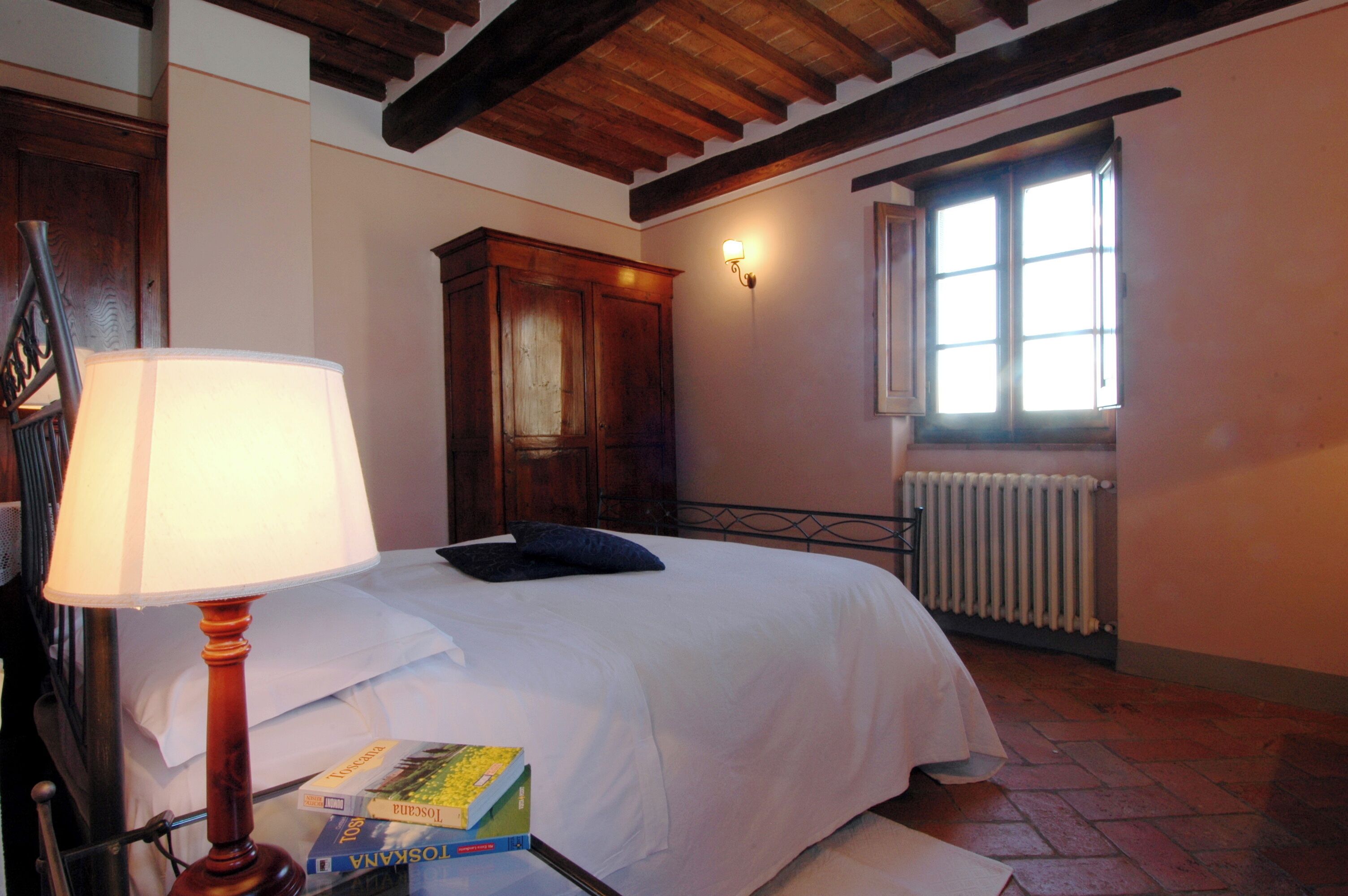 A bedroom with a bed, a nightstand with a lamp, a wooden cabinet, a radiator, and a window with natural light.
