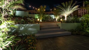 A nighttime outdoor area with steps leading to a covered seating area, surrounded by lush greenery and palm trees. - The Private Garden The Ambassador (Jan Thiel)