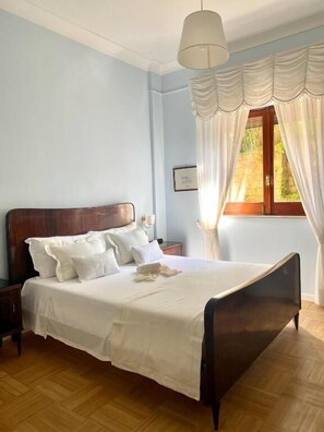 A bedroom with a wooden bed, white bedding, a nightstand, a bedside table, a window with curtains, and a hanging light fixture. - Maramà B&B Sorrento (Sorrento)