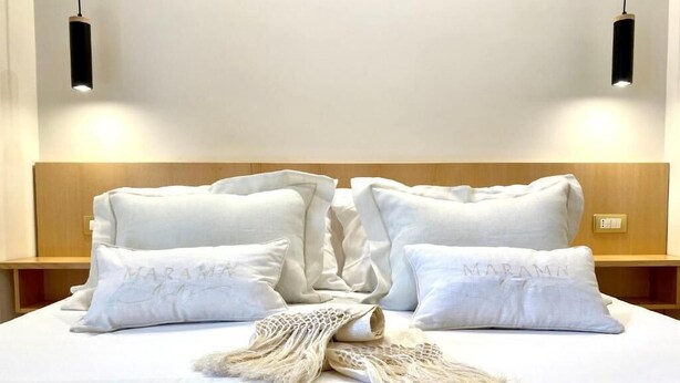 Una cama bien tendida con sábanas y almohadas blancas, un adorno de macramé y dos lámparas colgantes negras.