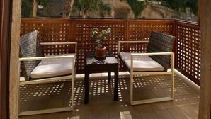 Un balcón con dos sillas de mimbre, una mesa, una valla de madera y vistas a árboles y edificios.