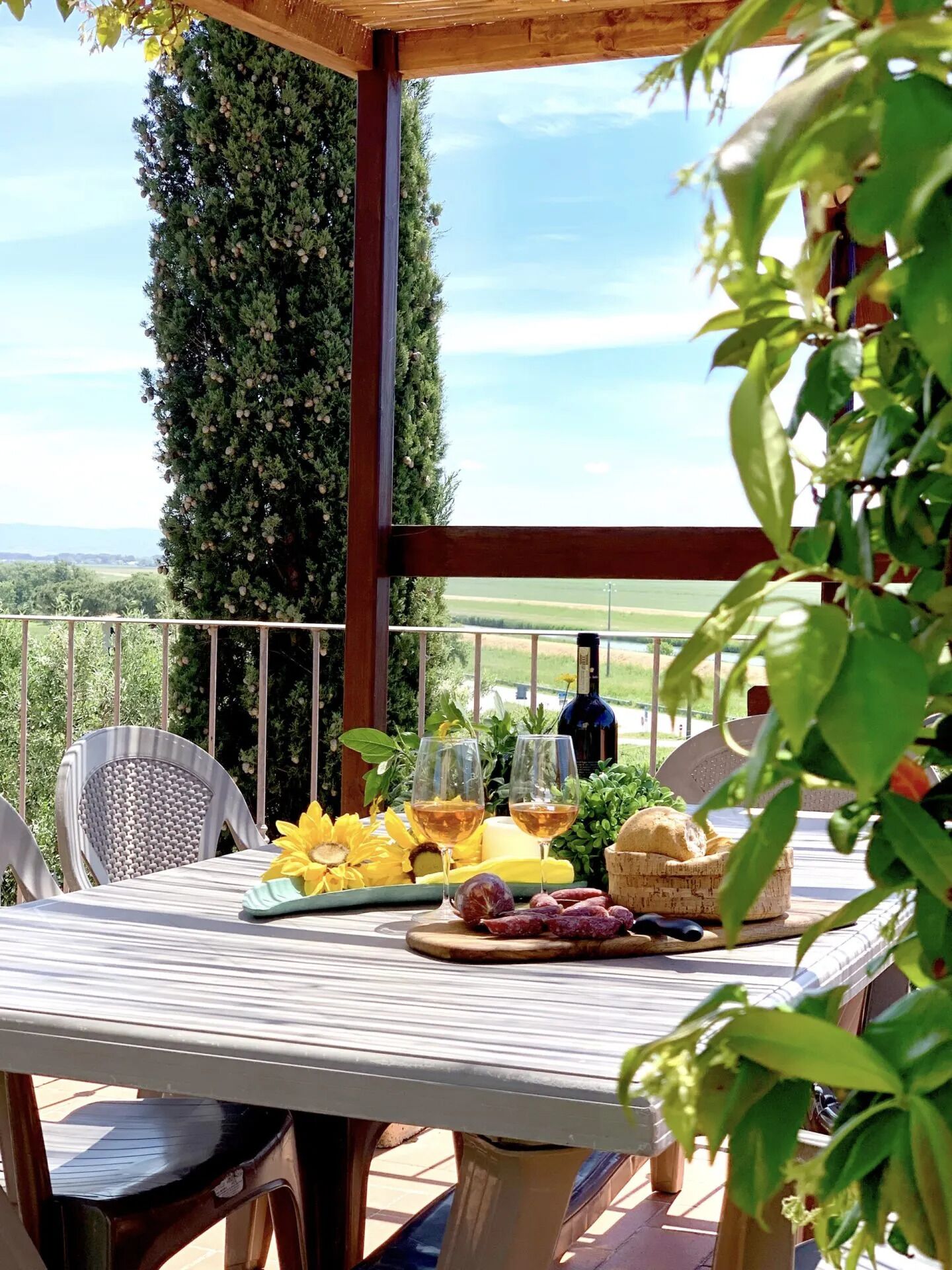 An outdoor dining area with a table set for two, featuring a bottle of wine, glasses, bread, and fruits.