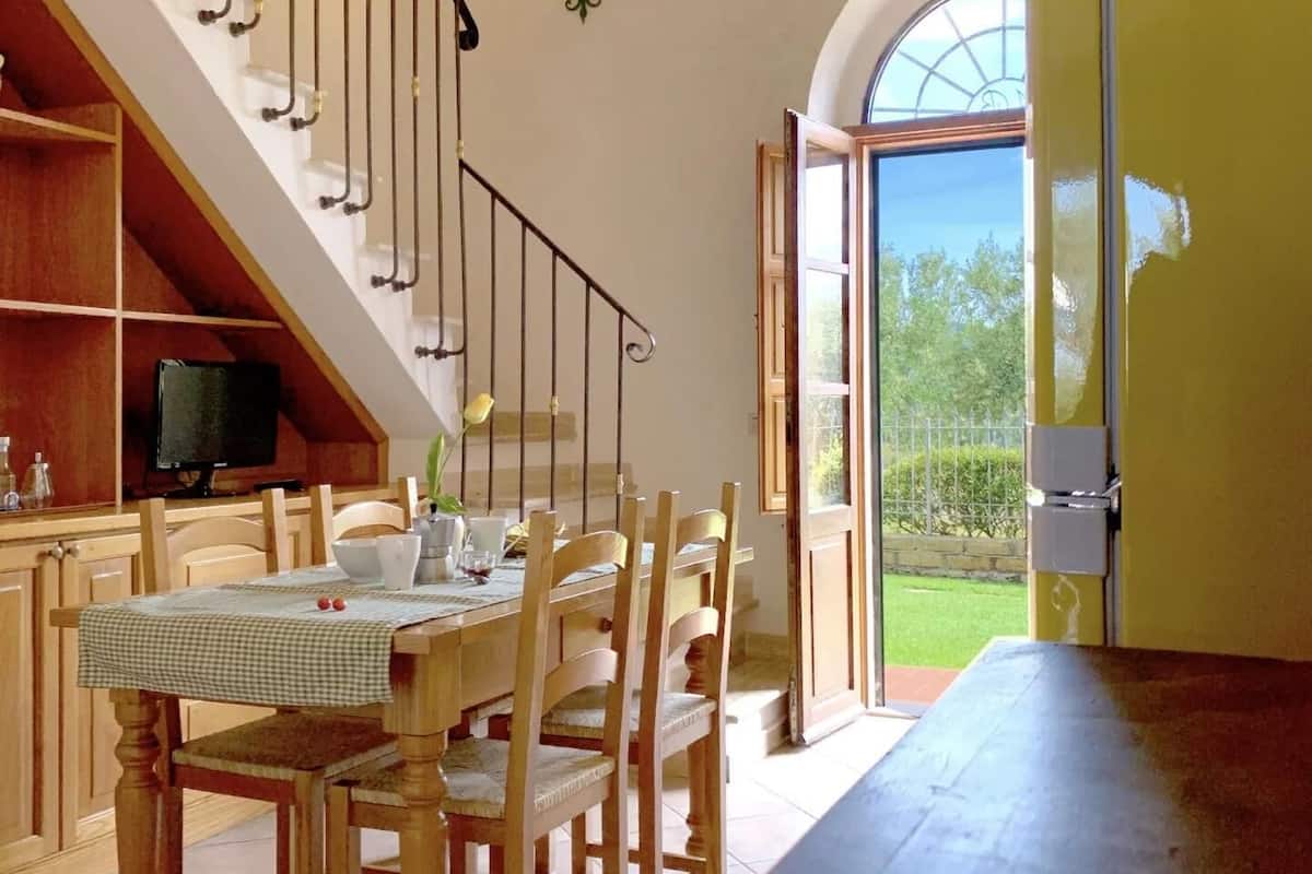 A dining area with a wooden table and chairs, a staircase with a TV in the background, and a view of the outdoors through an open door.