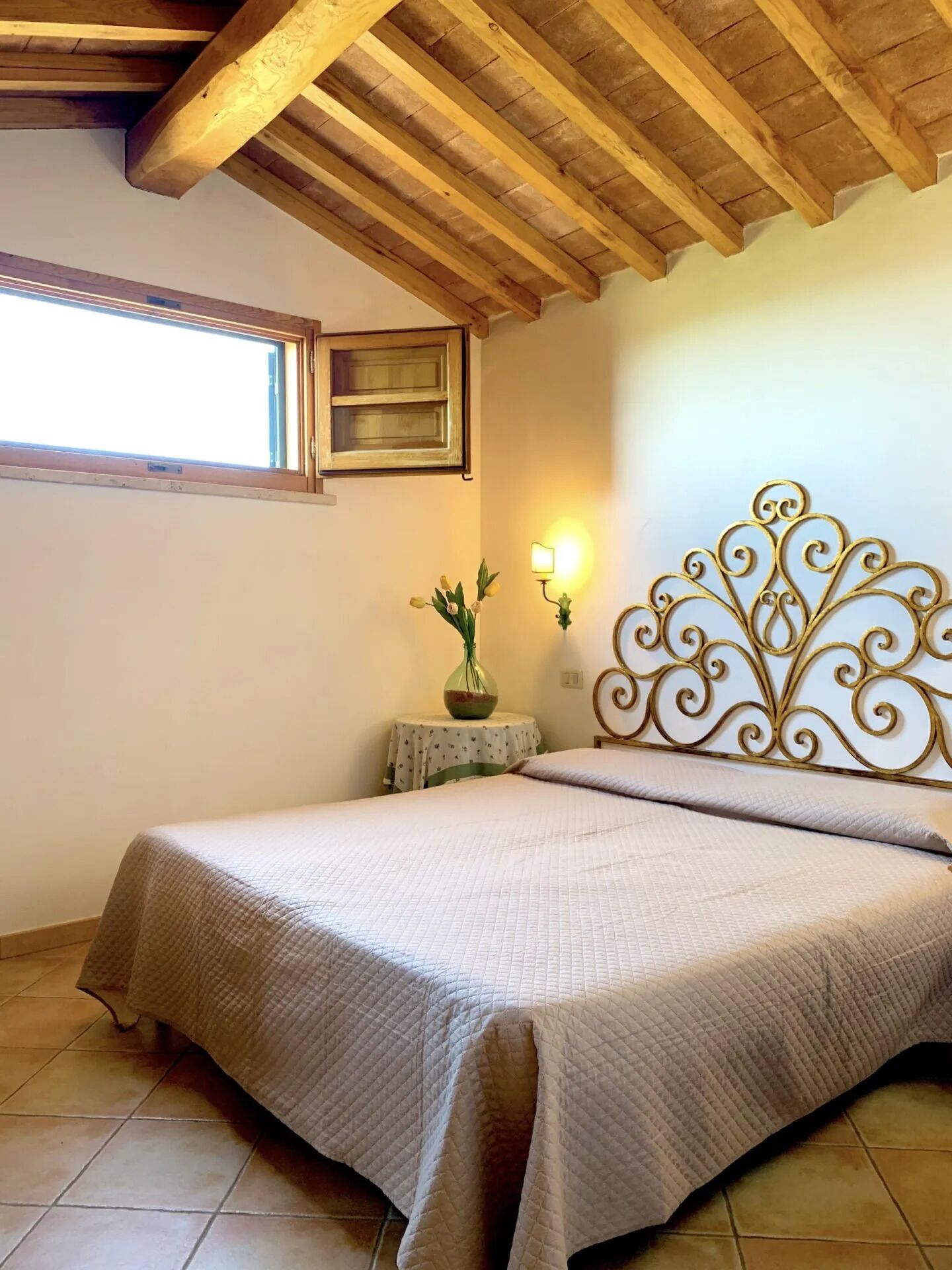 Une chambre avec un plafond en bois, un lit recouvert d’une courtepointe matelassée, une table de chevet ornée d’un vase de fleurs et un luminaire fixé au mur.
