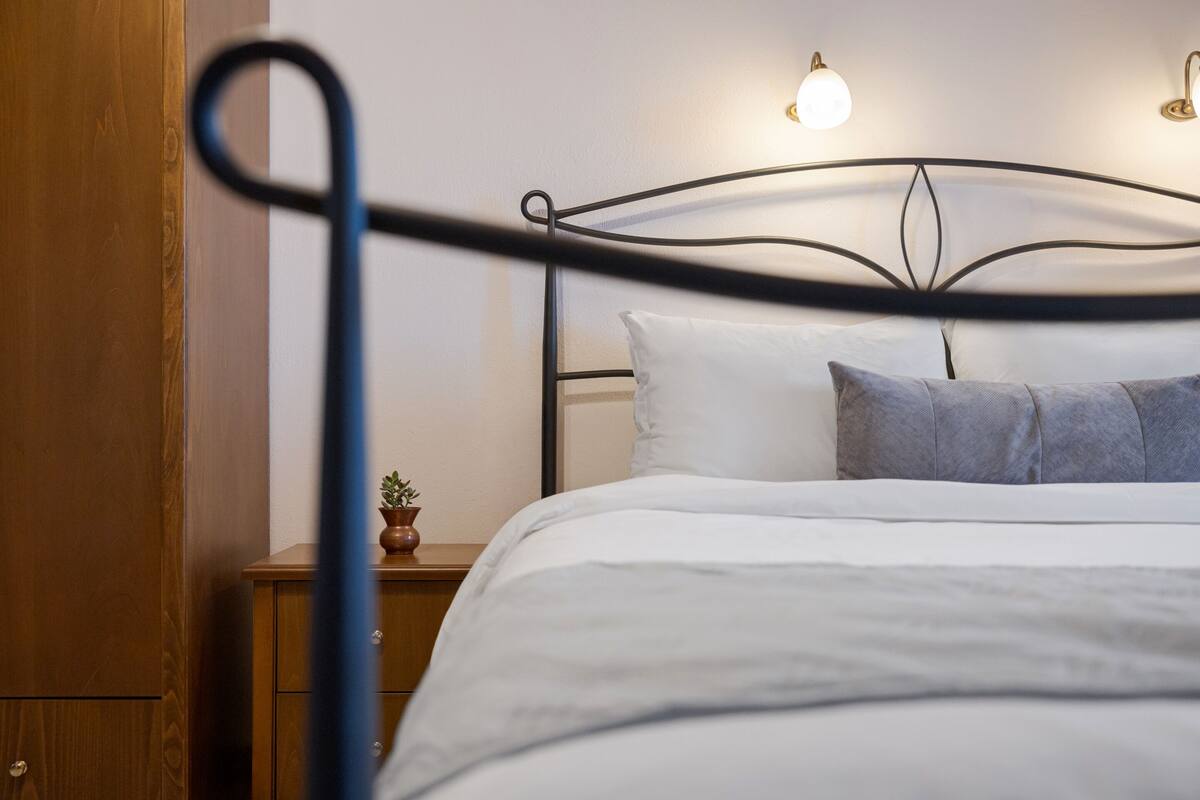Ein ordentlich bezogenes Bett mit weißer und grauer Bettwäsche, ein Nachttisch aus Holz mit einer kleinen Pflanze und ein Metall-Kopfteil mit an der Wand befestigten Lampen.