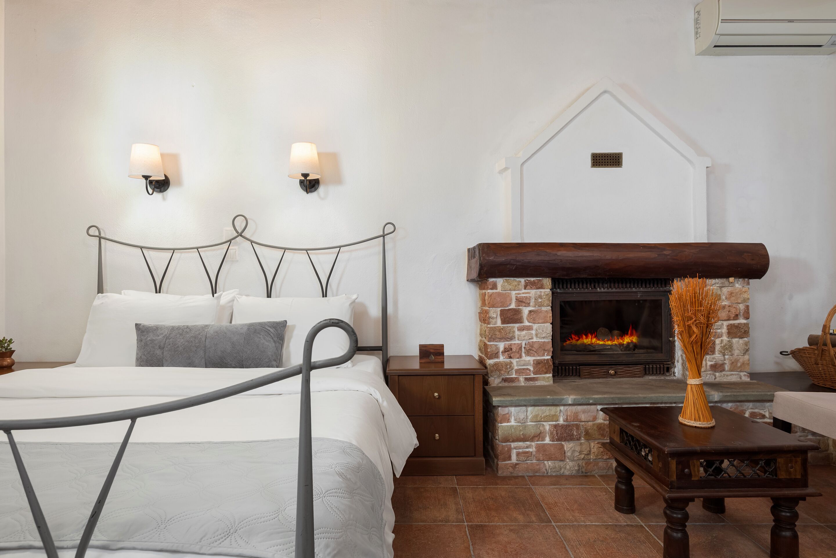 A bedroom with a metal bed frame, white bedding, a nightstand, a wooden table with a vase, and a stone fireplace with a fire.