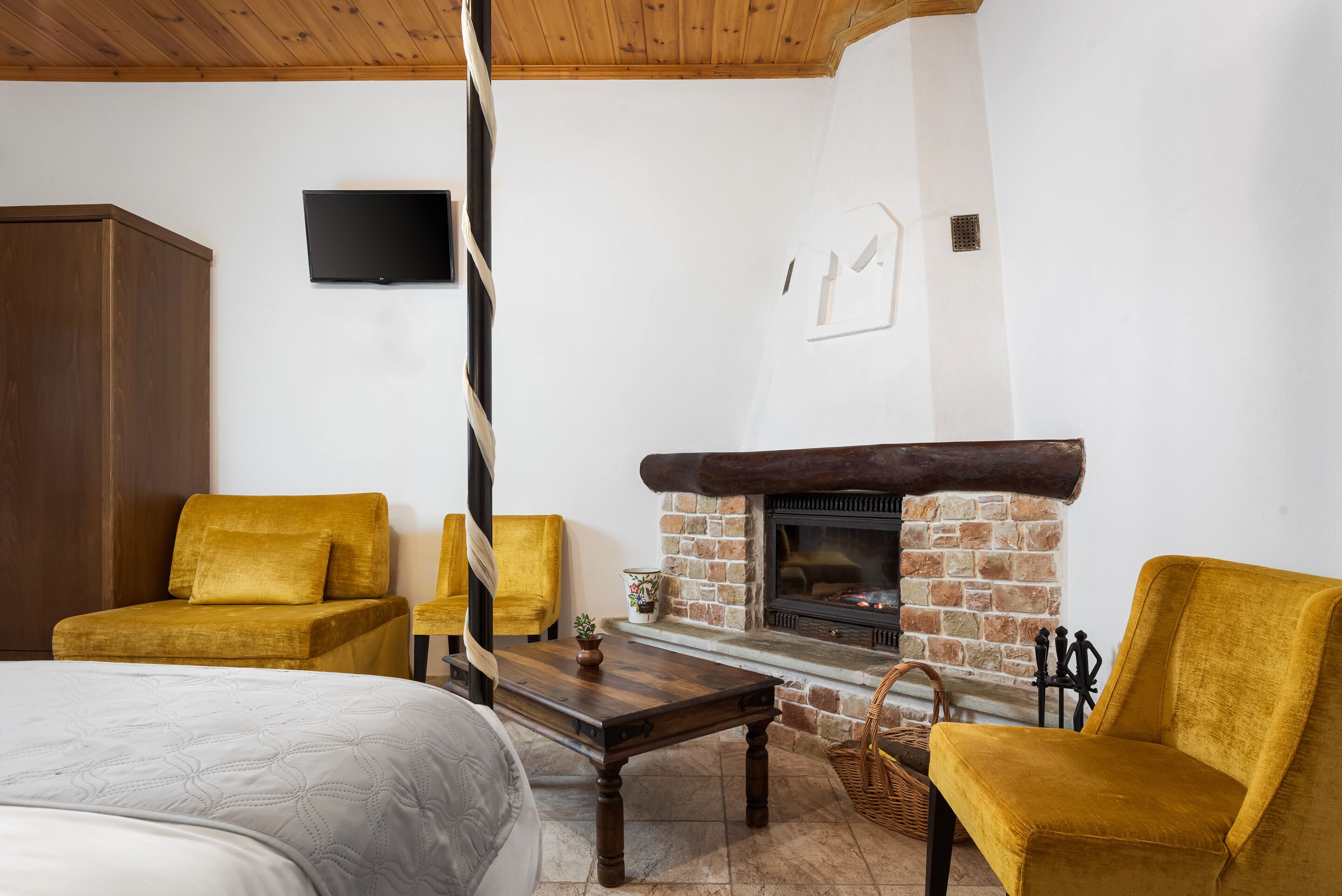 Una habitación acogedora con una cama, dos sillones amarillos, una mesa de madera, una chimenea y un televisor montado en la pared.