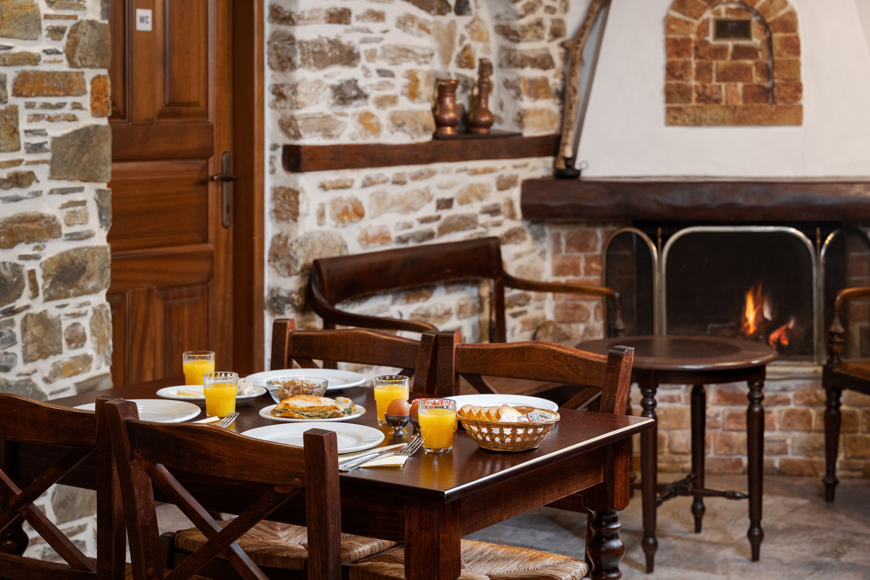 A dining table set with plates, glasses, and a basket of bread, surrounded by wooden chairs in a stone-walled room with a fireplace.