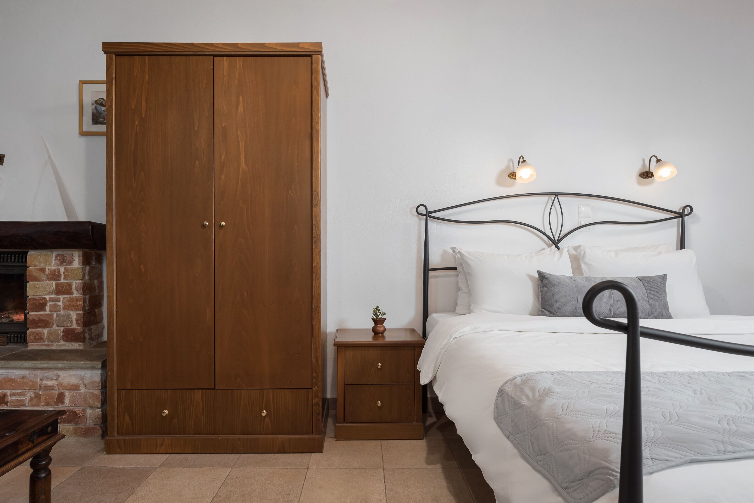 A bedroom with a wooden wardrobe, a metal bed frame, a bedside table with a lamp, and a small plant.