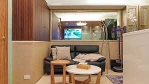 Ein modernes Wohnzimmer mit einem schwarzen Ledersofa, zwei Holfnitztischen, einem weißen Couchtisch und einem großen Spiegel, der einen Fernsehbildschirm reflektiert.
