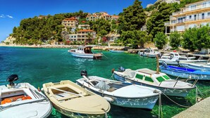 A marina with various boats docked, a clear blue sea, and buildings with balconies on a hillside.