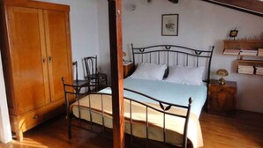 A bedroom with a wooden bed frame, a single bed, a wooden wardrobe, a chair, a bedside table with a lamp, and a wall-mounted shelf with books.