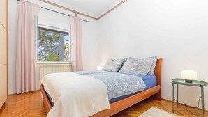 A bedroom with a wooden bed, a window with curtains, a bedside table with a lamp, and a radiator. - Holiday apartment mit Klimaanlage (Pula)
