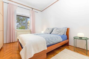 A bedroom with a wooden bed, a window with curtains, a bedside table with a lamp, and a radiator. - Holiday apartment mit Klimaanlage (Pula)