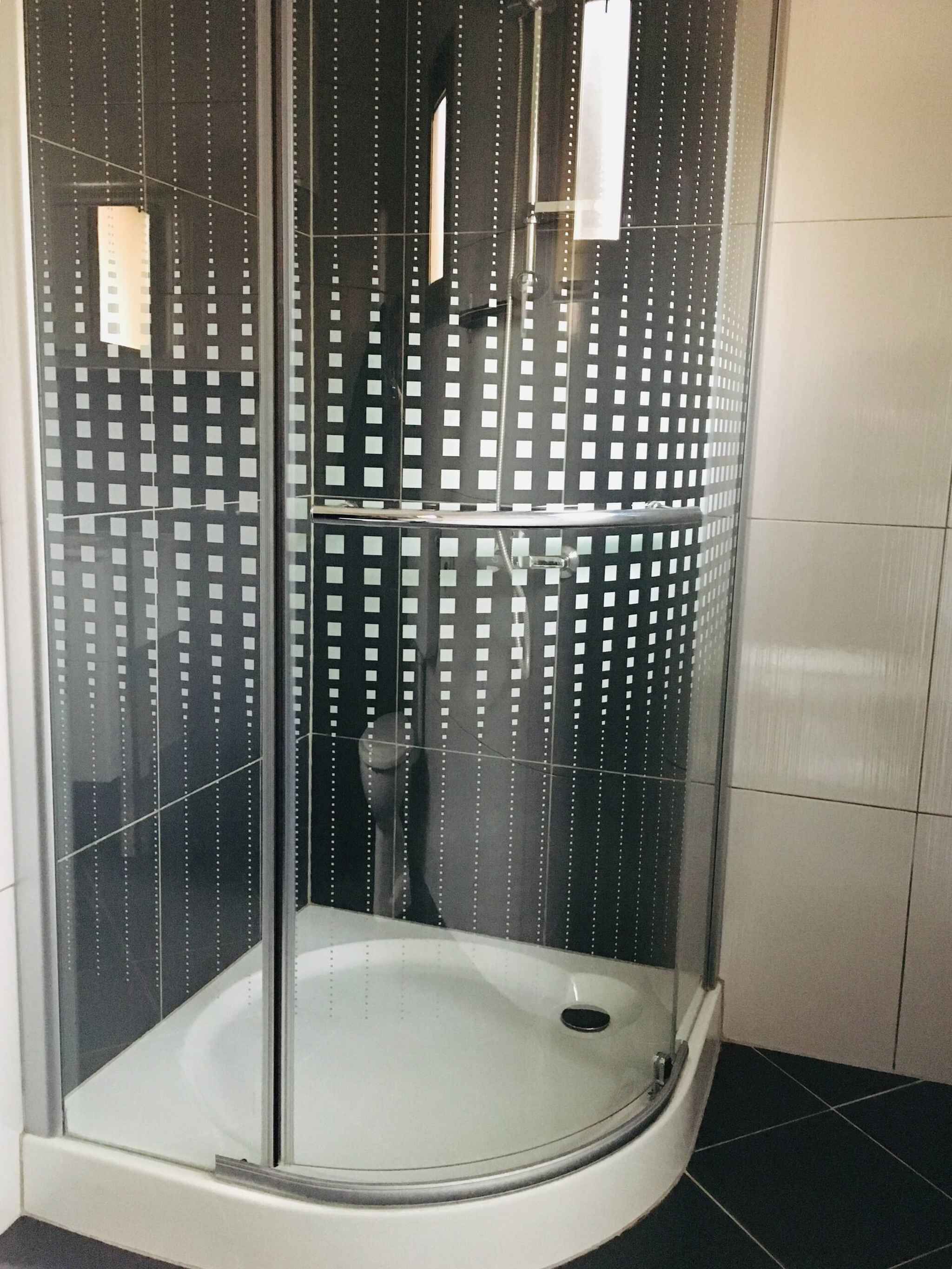A glass-enclosed shower stall with a white floor, black tiled walls, and a visible drain.