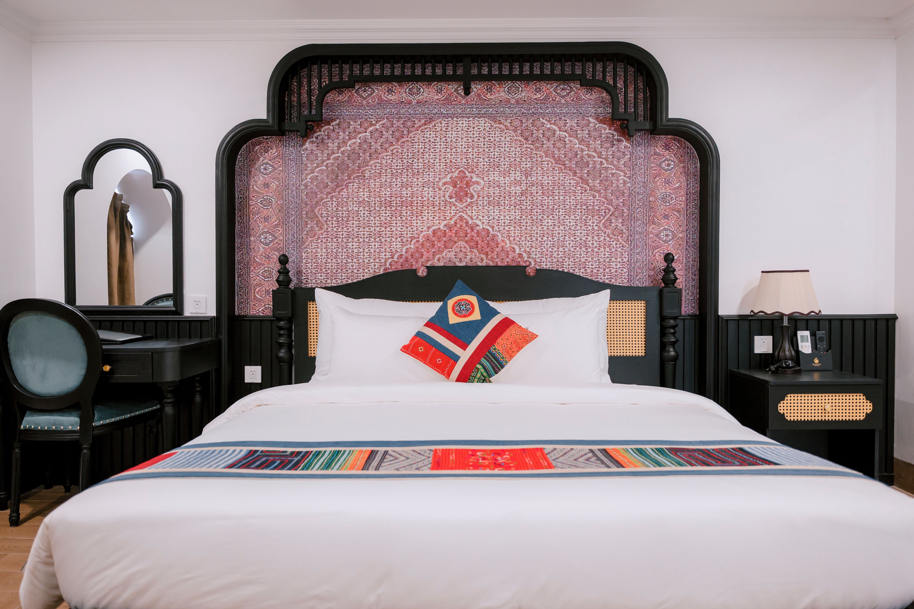A neatly made bed with a patterned bedspread, a headboard with a detailed design, a bedside table with a lamp, and a chair with a mirror.