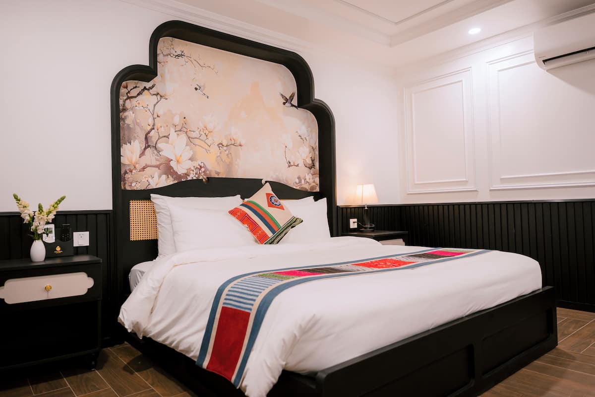 Una cama bien hecha con sábanas blancas y un colorido plaid, un cabecero negro con diseño floral, una mesita de noche con lámpara y una obra de arte colgada en la pared.