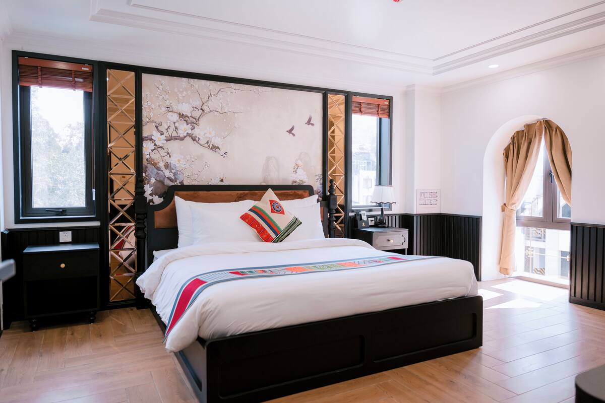 Un dormitorio ordenado con una cama grande, una mesita de noche, un televisor instalado en la pared y una ventana con cortinas.