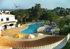 A swimming pool with sun loungers and umbrellas, surrounded by palm trees and a building with a tiled roof. - Villa with air conditioning, balcony and pool (Porches)