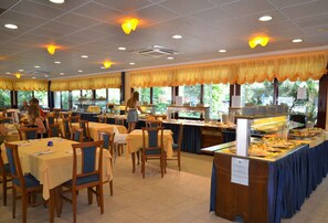 A buffet-style dining area with multiple tables and chairs, a buffet counter with various food options, and large windows allowing natural light. - Holiday apartment with outdoor pool (Bibione)