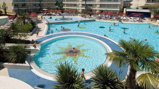 Una amplia piscina al aire libre rodeada de palmeras y tumbonas, con varios edificios de hotel al fondo.