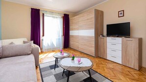 Ein modernes Wohnzimmer mit einer Couch, zwei kleinen Tischen mit Dekoration, einem Fernseher auf einer Kommode und einem großen Fenster mit Vorhängen.