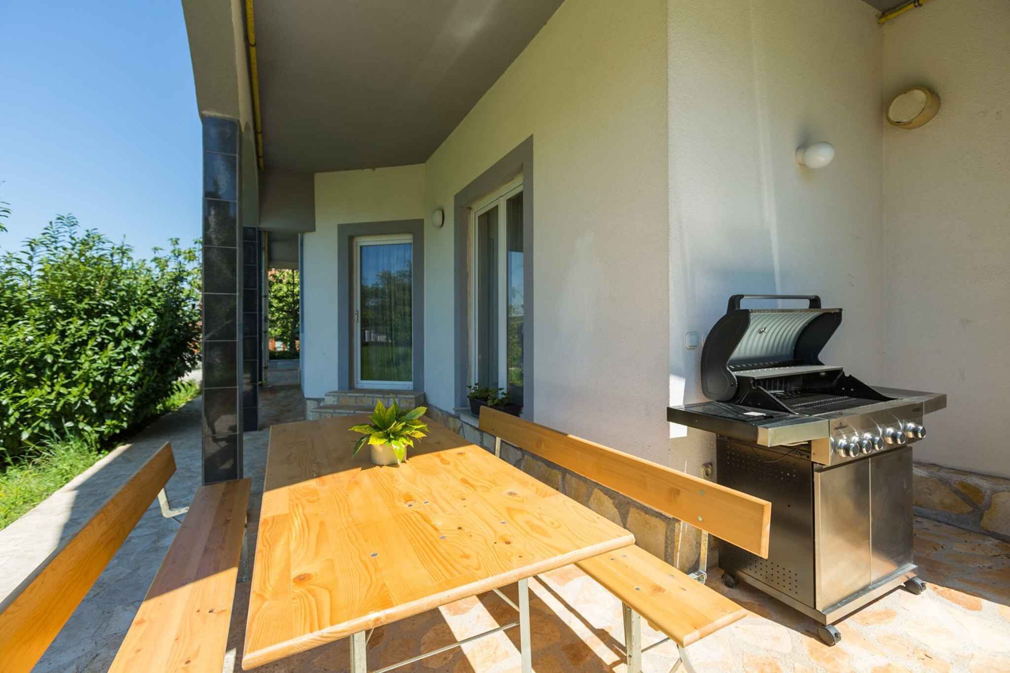 An outdoor patio with a wooden table and benches, a stainless steel grill, and a view of a house in the background.