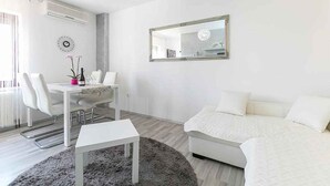 Ein modernes, minimalistisches Zimmer mit einem weißen Sofa, einem Esstisch mit Stühlen, einem kleinen weißen Tisch und einem großen Spiegel an der Wand.