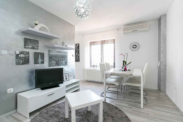 Una sala de estar moderna con televisor de pantalla plana, muebles blancos, un comedor con mesa y sillas, y obras de arte en las paredes.