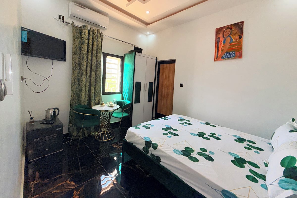 A hotel room with a bed, a desk with a chair, a TV mounted on the wall, and a window with curtains.