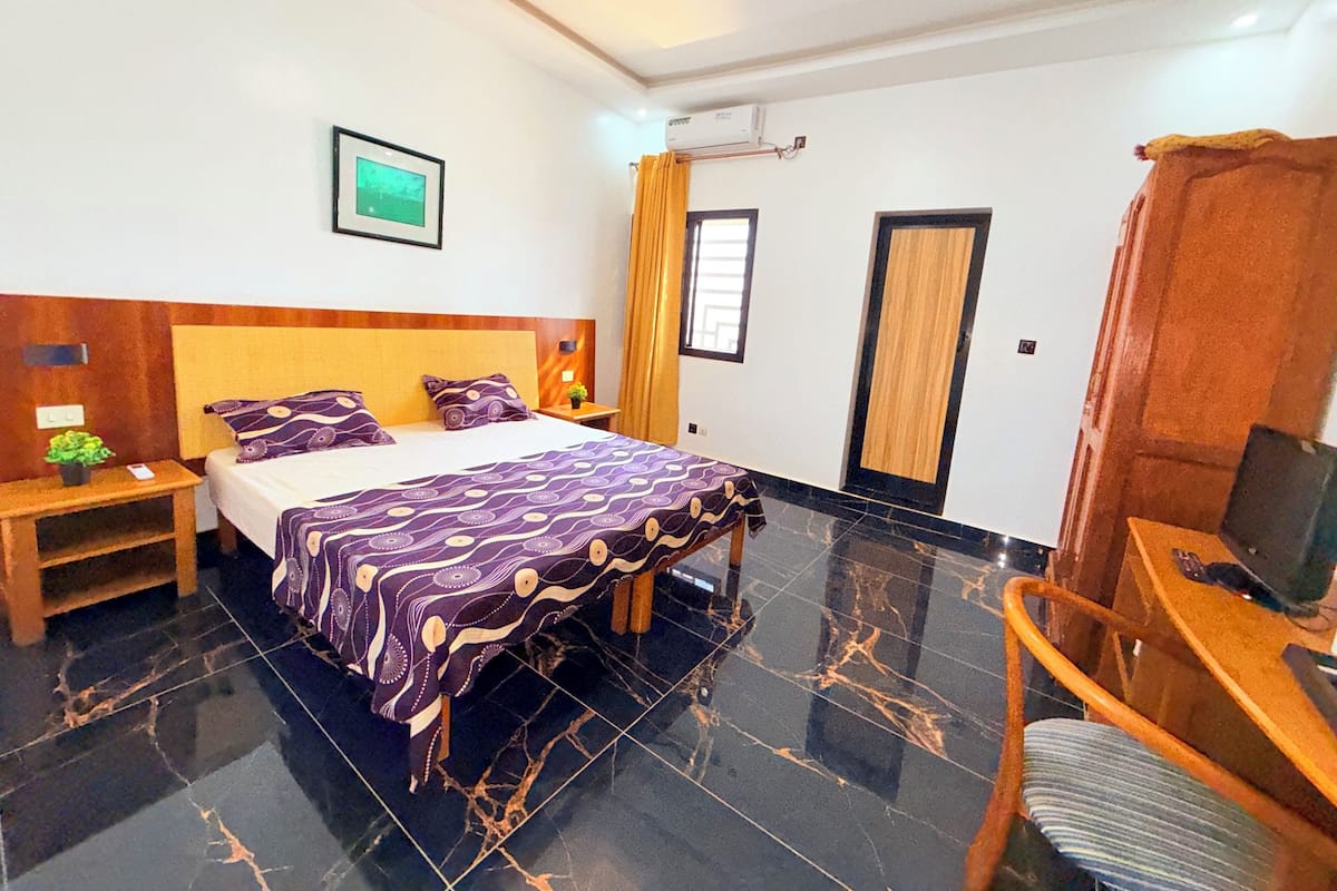 A hotel room with a bed, a desk with a chair, a nightstand, a wall-mounted TV, and a window with curtains.