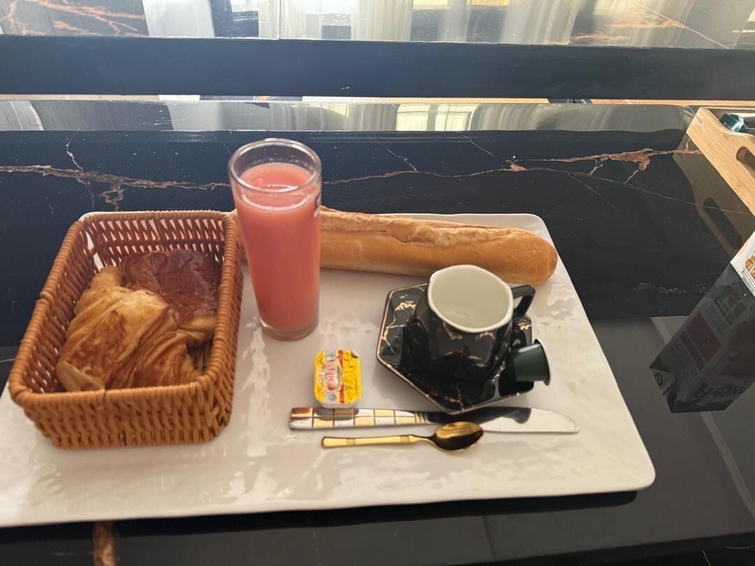 A breakfast tray with a basket of pastries, a glass of juice, a baguette, a cup, a small container of jam, and cutlery on a marble table.