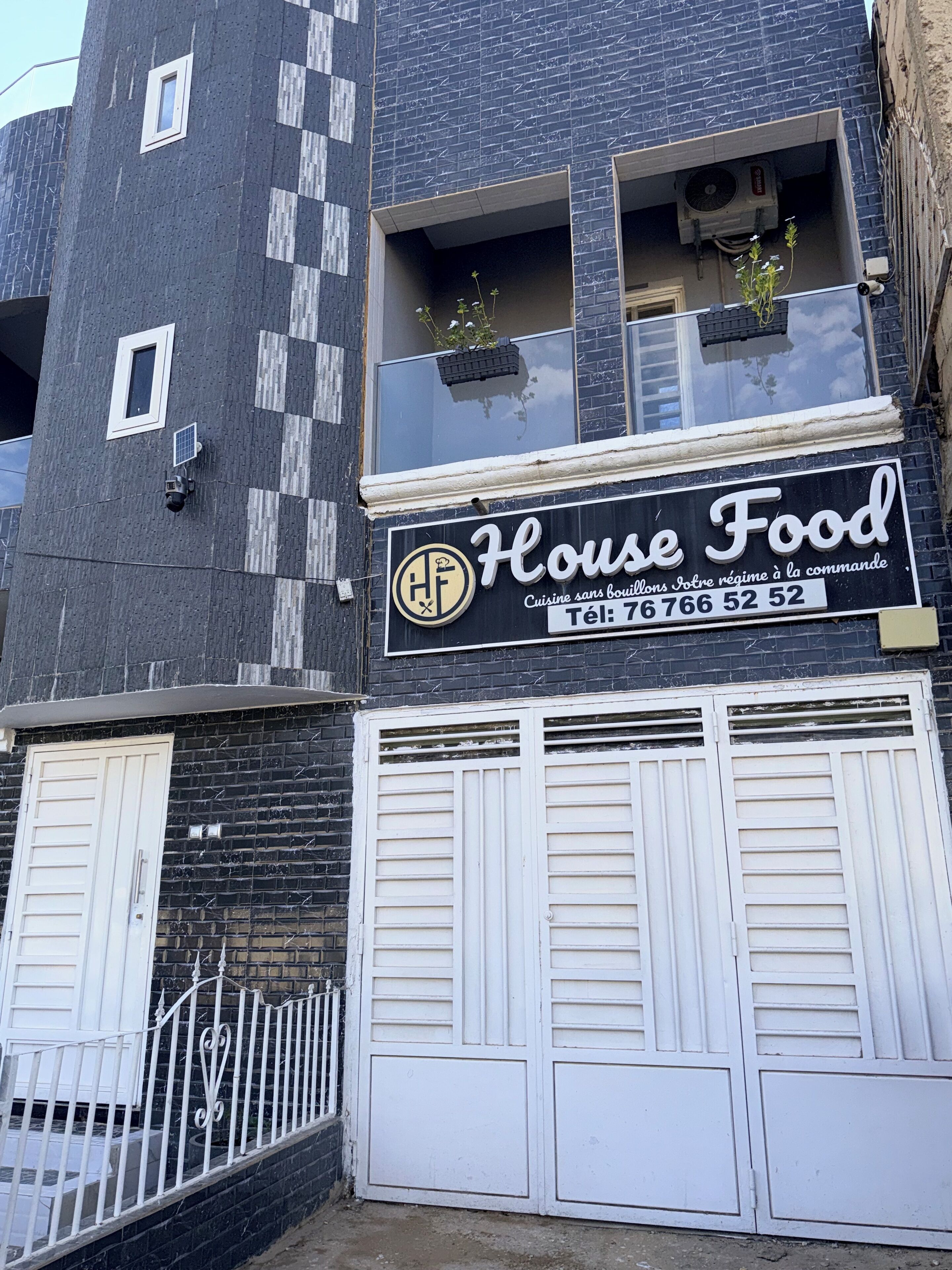 A building with a sign reading "House Food" and a phone number. There are two windows with plants, a balcony, and a white door.