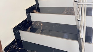 Una escalera con escalones de mármol blanco y negro, barandilla de metal y una pared blanca con dos interruptores de luz.