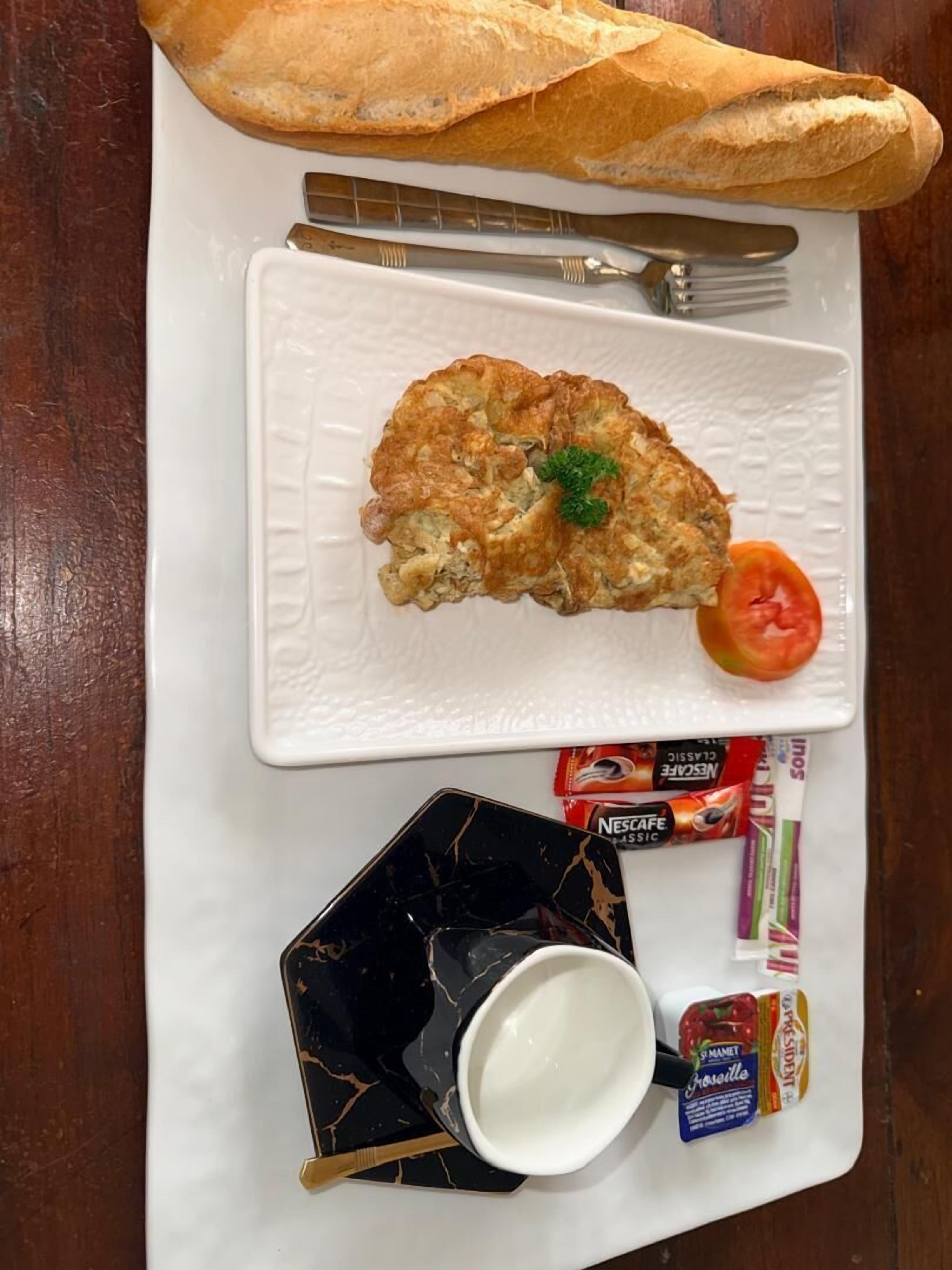 A meal with a croissant, a tomato, and a cup of coffee on a white plate, with a baguette and utensils on a wooden table.