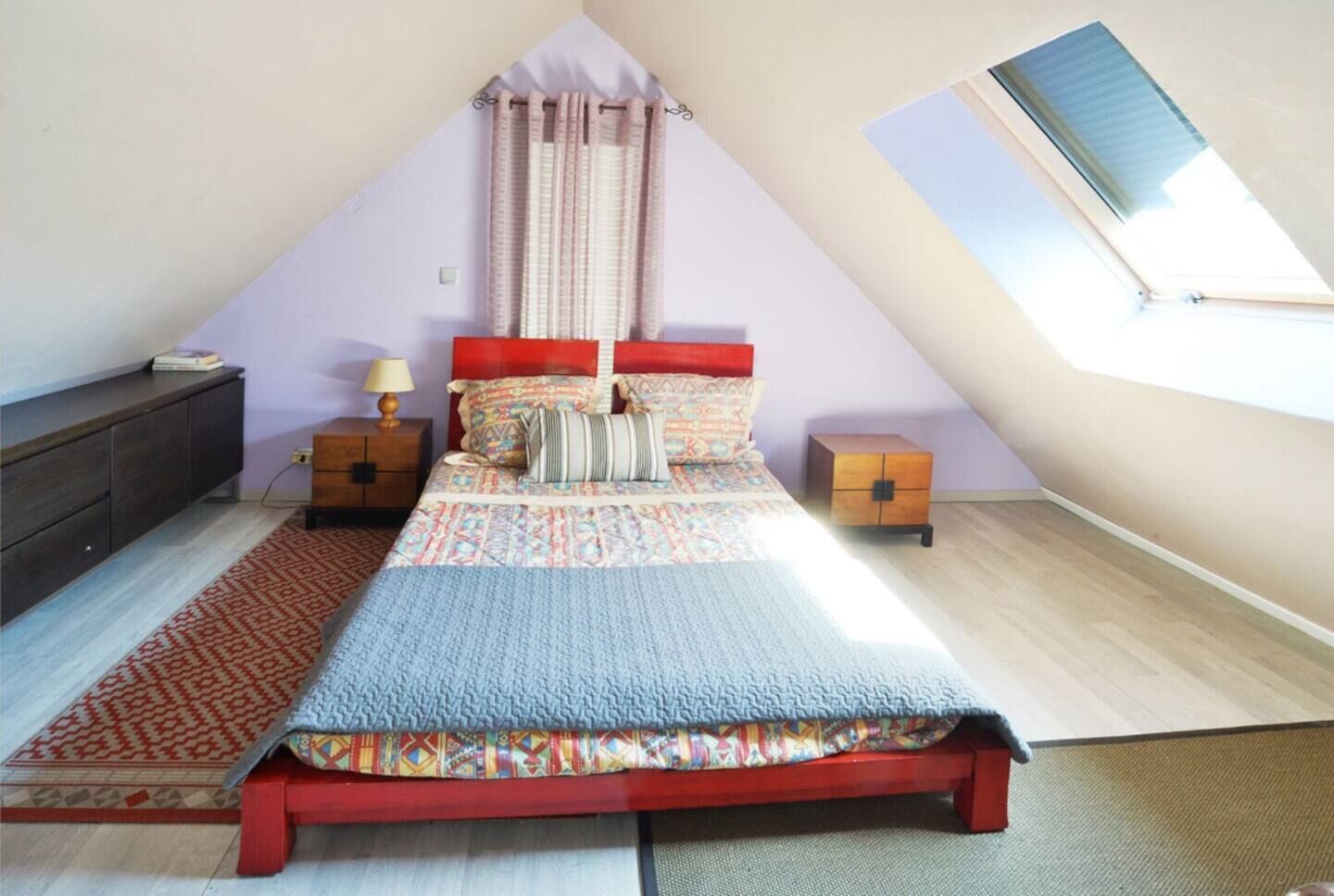 A bedroom with a sloped ceiling, a bed with a red headboard, a nightstand with a lamp, a small wooden chest, and a red patterned rug.