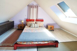 A bedroom with a sloped ceiling, a bed with a red headboard, a nightstand with a lamp, a small wooden chest, and a red patterned rug. - Small house (Férolles-Attilly)