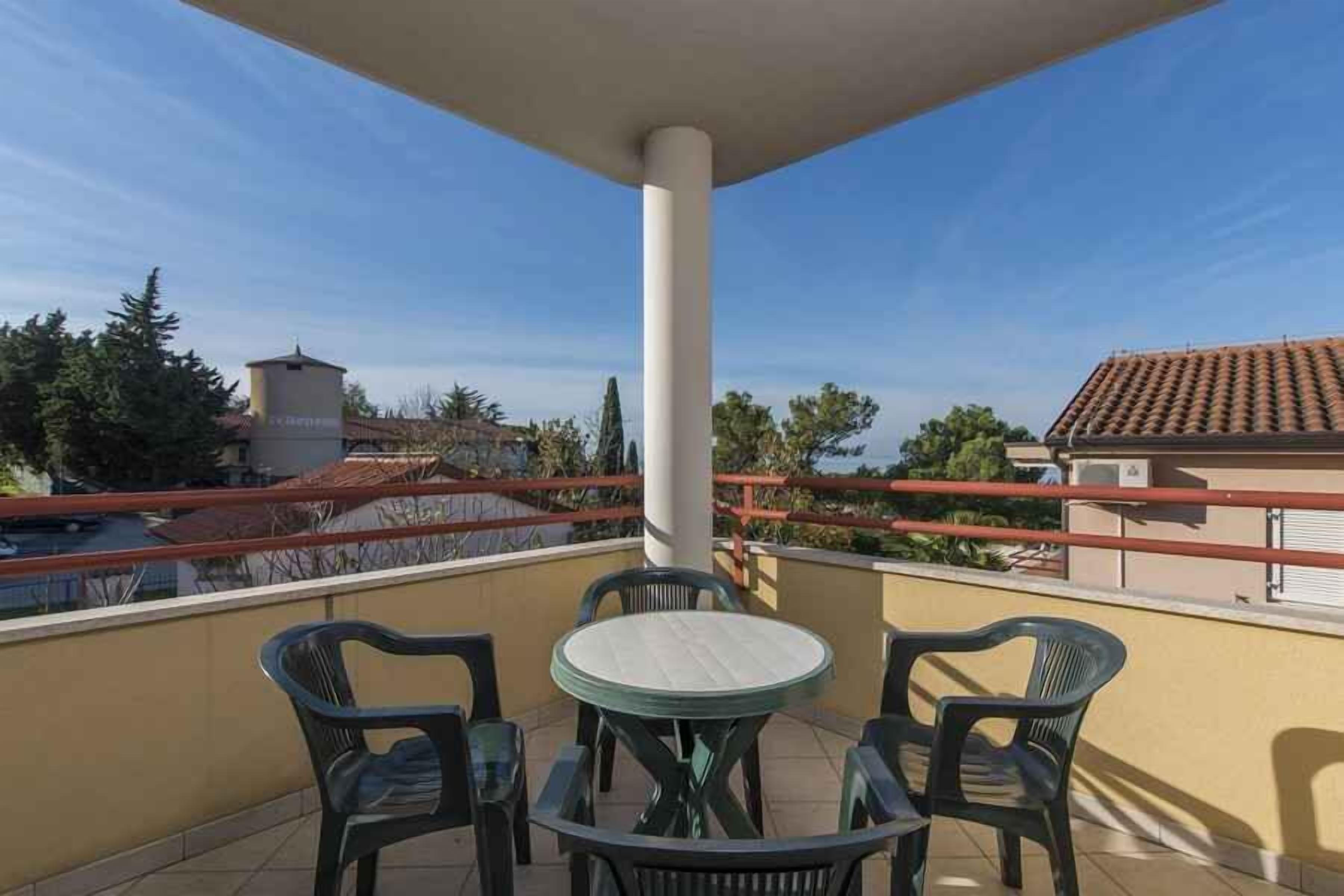 A balcony with a round table and four chairs, a white pillar, and a view of neighboring buildings and trees.