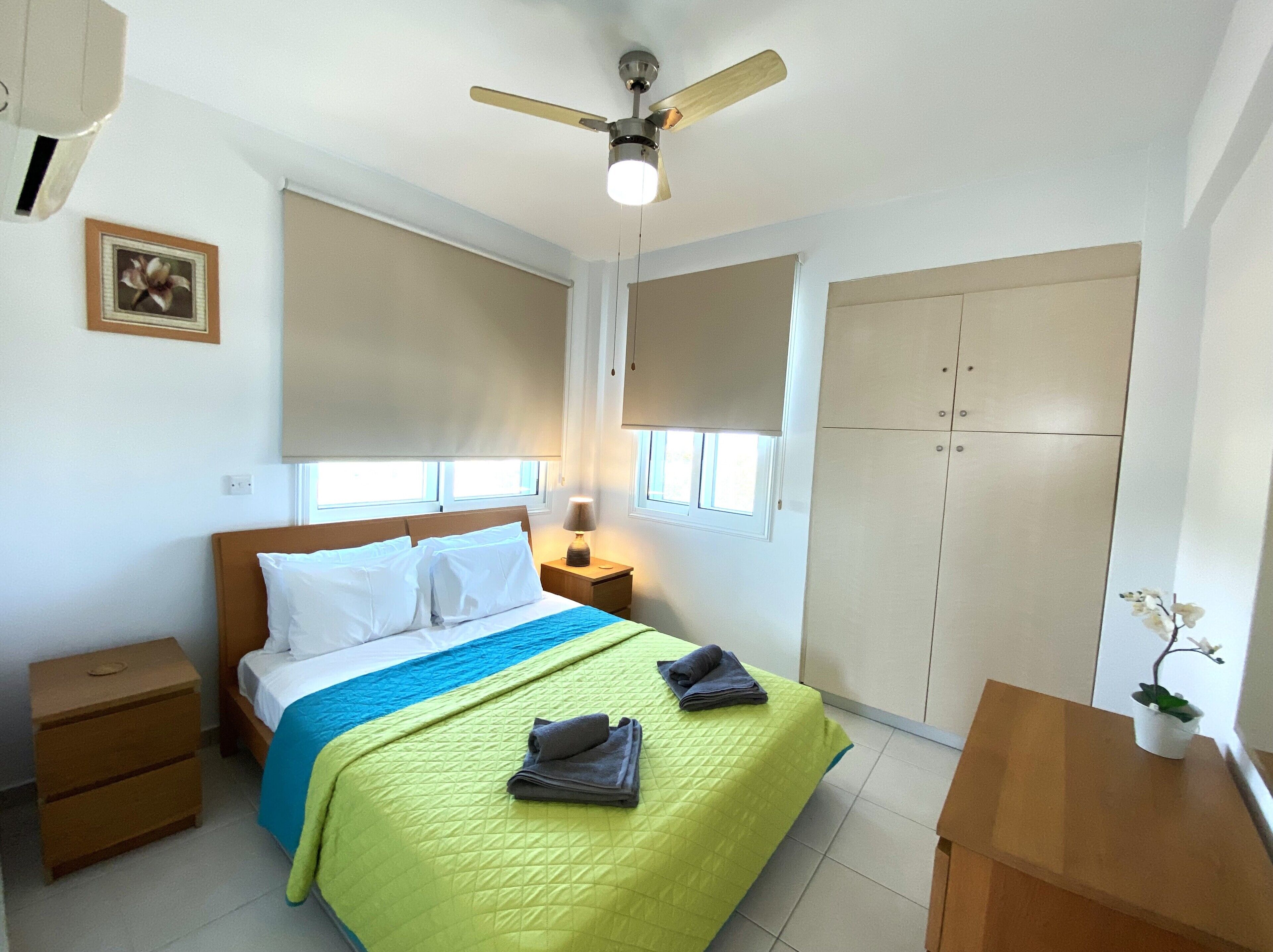 Ein Schlafzimmer mit einem Bett, einem Nachttisch, einer Nachttischlampe, einem Deckenventilator, einem Kleiderschrank und einem kleinen Tisch mit einem Topfplanzen.