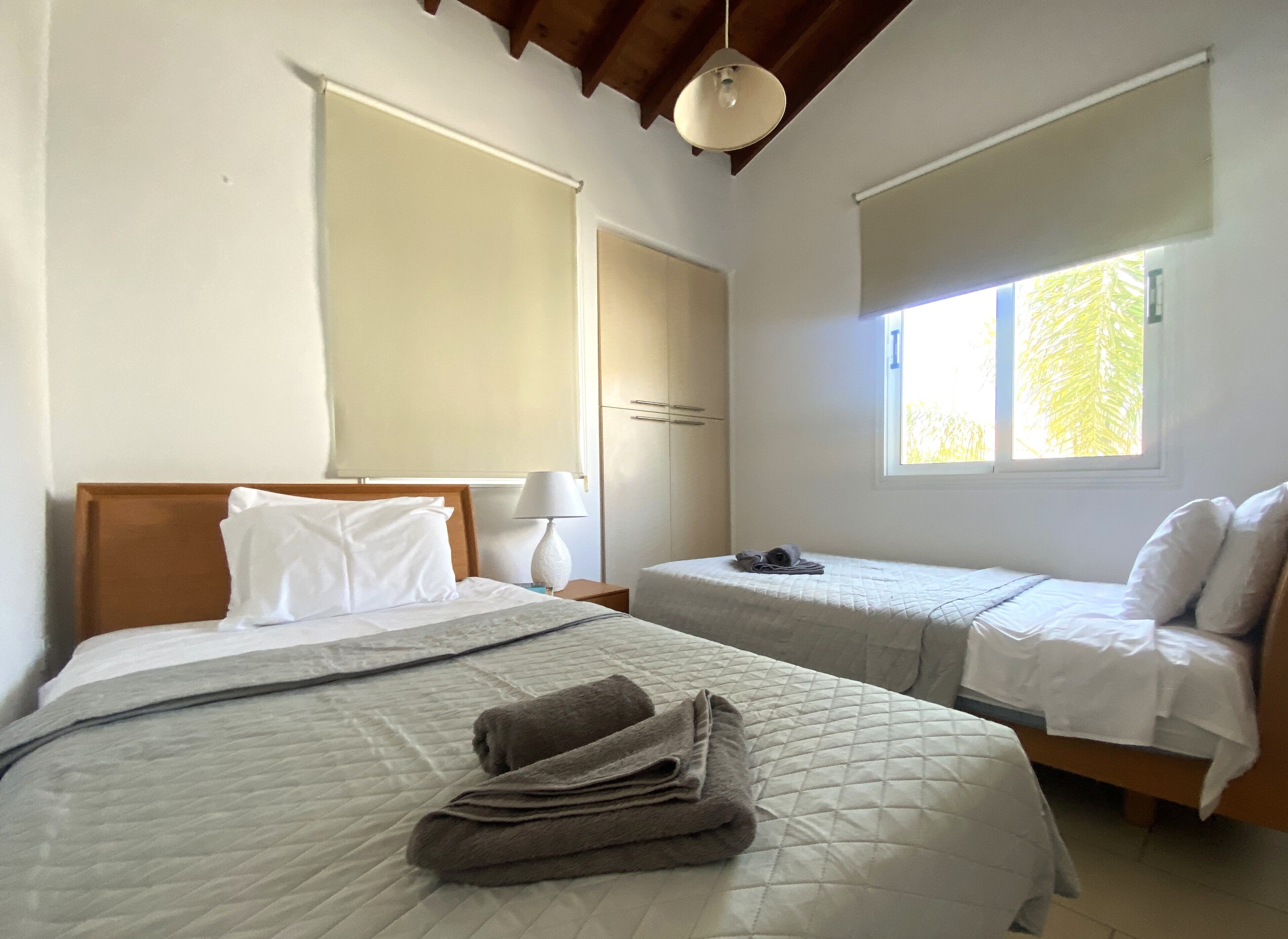 Ein Schlafzimmer mit zwei Betten, einem Fenster mit Jalousien, einer Lampe und einem Deckenventilator.