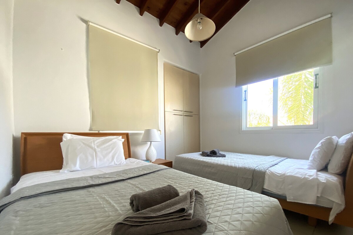 Ein Schlafzimmer mit zwei Betten, einem Fenster mit Jalousien, einer Lampe und einem Deckenventilator.