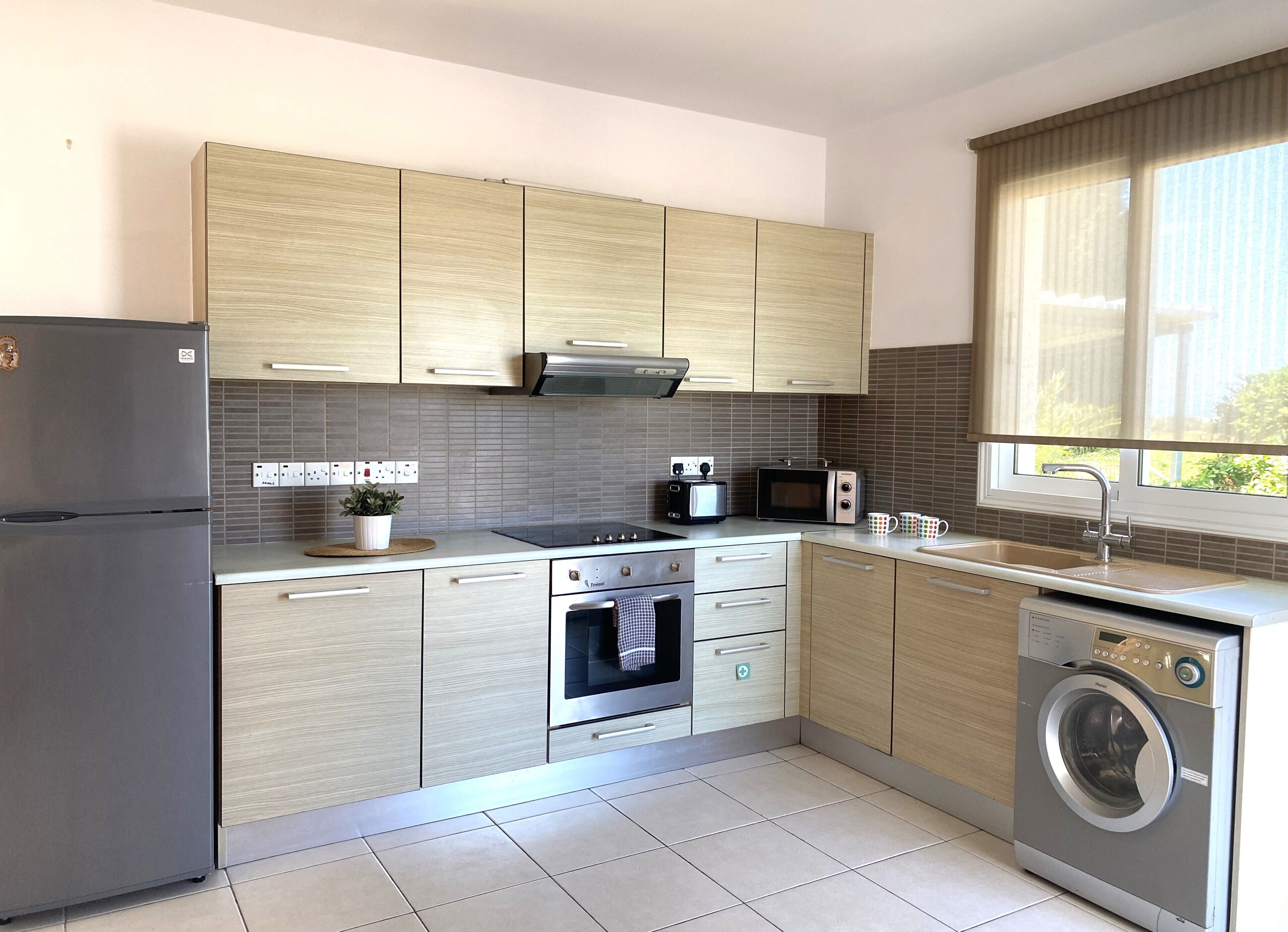 A modern kitchen with a built-in oven, microwave, and washing machine, light wood cabinets, and a window with curtains.