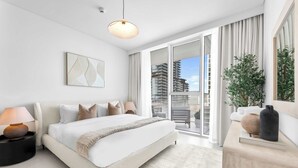 Une chambre moderne avec un grand lit, une table de chevet avec une lampe, une grande fenêtre donnant sur des immeubles et une plante posée sur une petite table d’appoint.