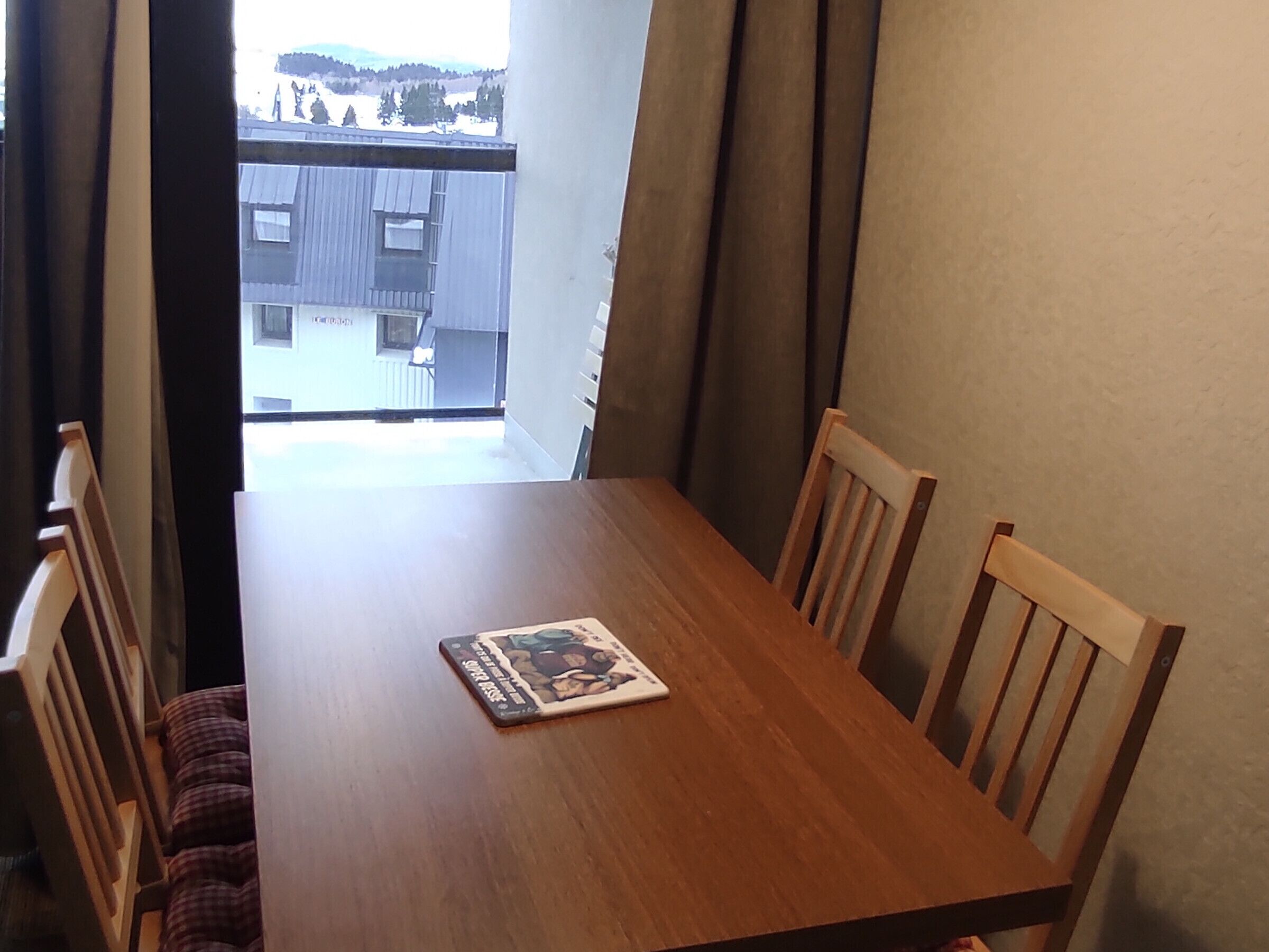 Une table de salle à manger en bois, quatre chaises, un livre posé sur la table et, par la fenêtre, une vue sur un bâtiment derrière des rideaux.